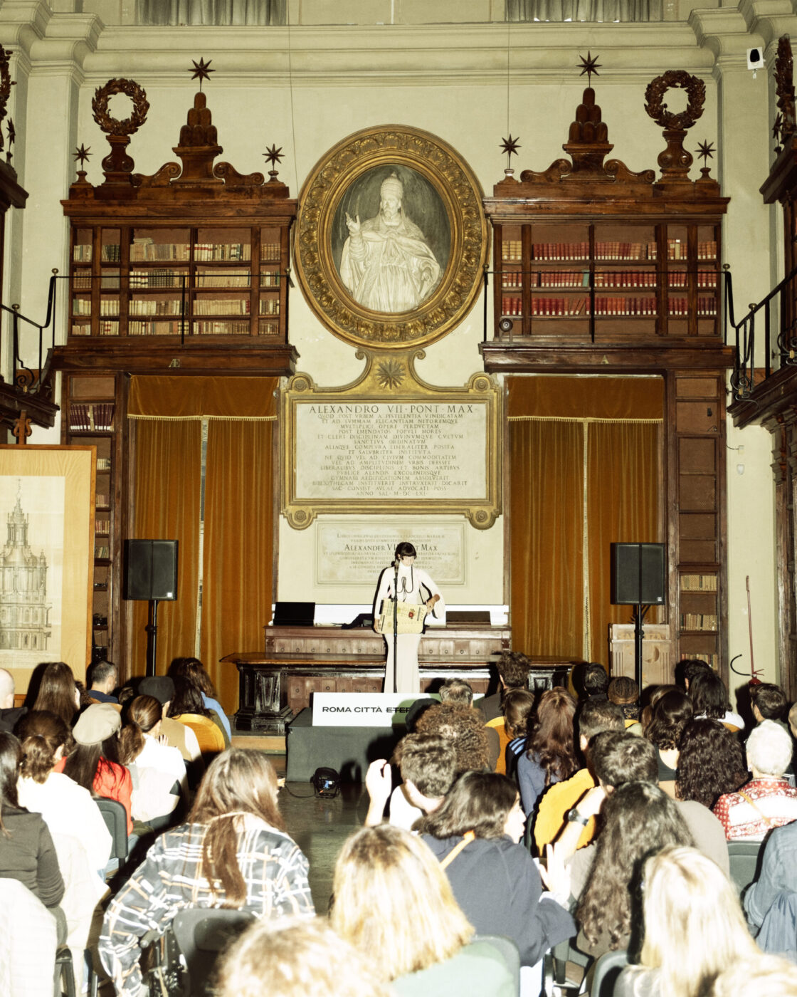 A speaker at a podium addresses an audience in a stately library with tall shelves and ornate decor; people sit facing forward.