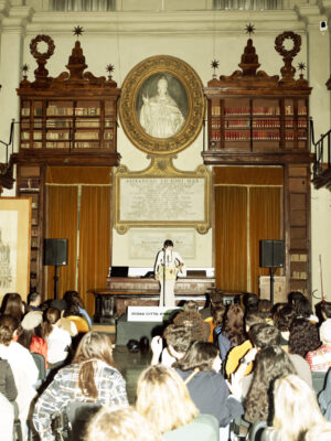 A speaker at a podium addresses an audience in a stately library with tall shelves and ornate decor; people sit facing forward.