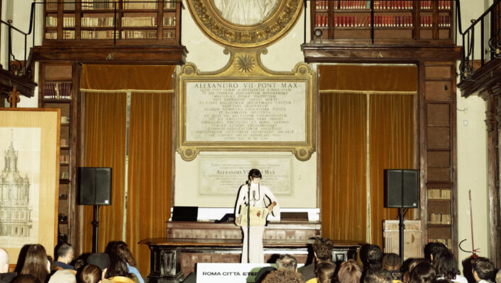 A speaker at a podium addresses an audience in a stately library with tall shelves and ornate decor; people sit facing forward.