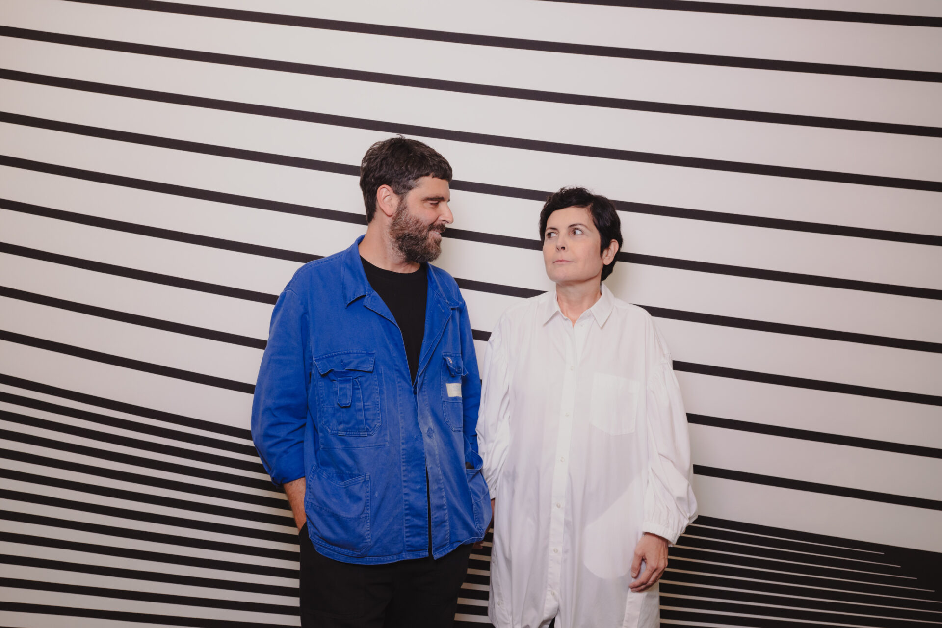 A man in a blue jacket and a woman in a white shirt face each other before a wall with black diagonal stripes, both expressionless.