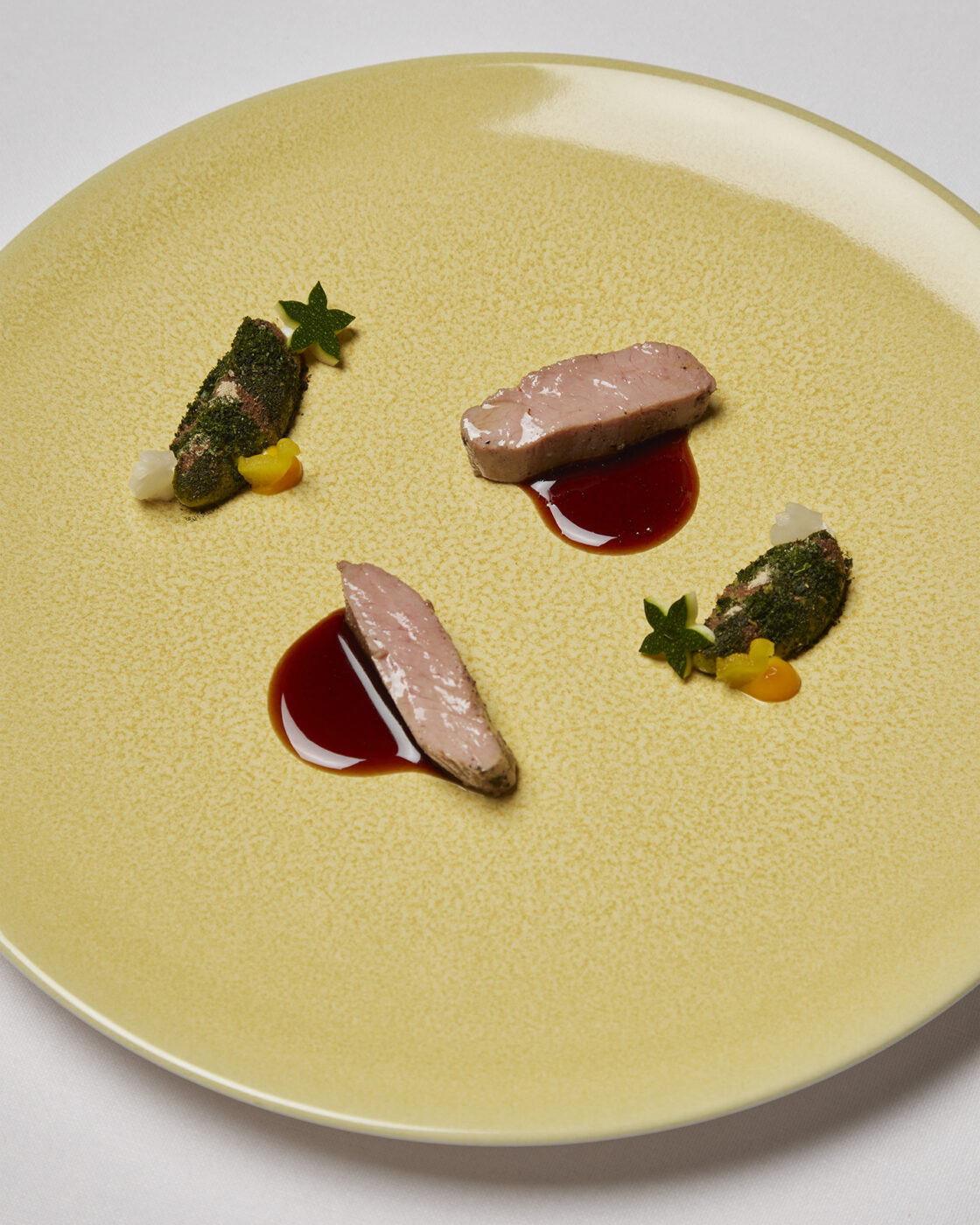 A yellow plate holds three seared meat slices with dark sauce, green herbs, yellow dollops, and white cubes on a white tablecloth.