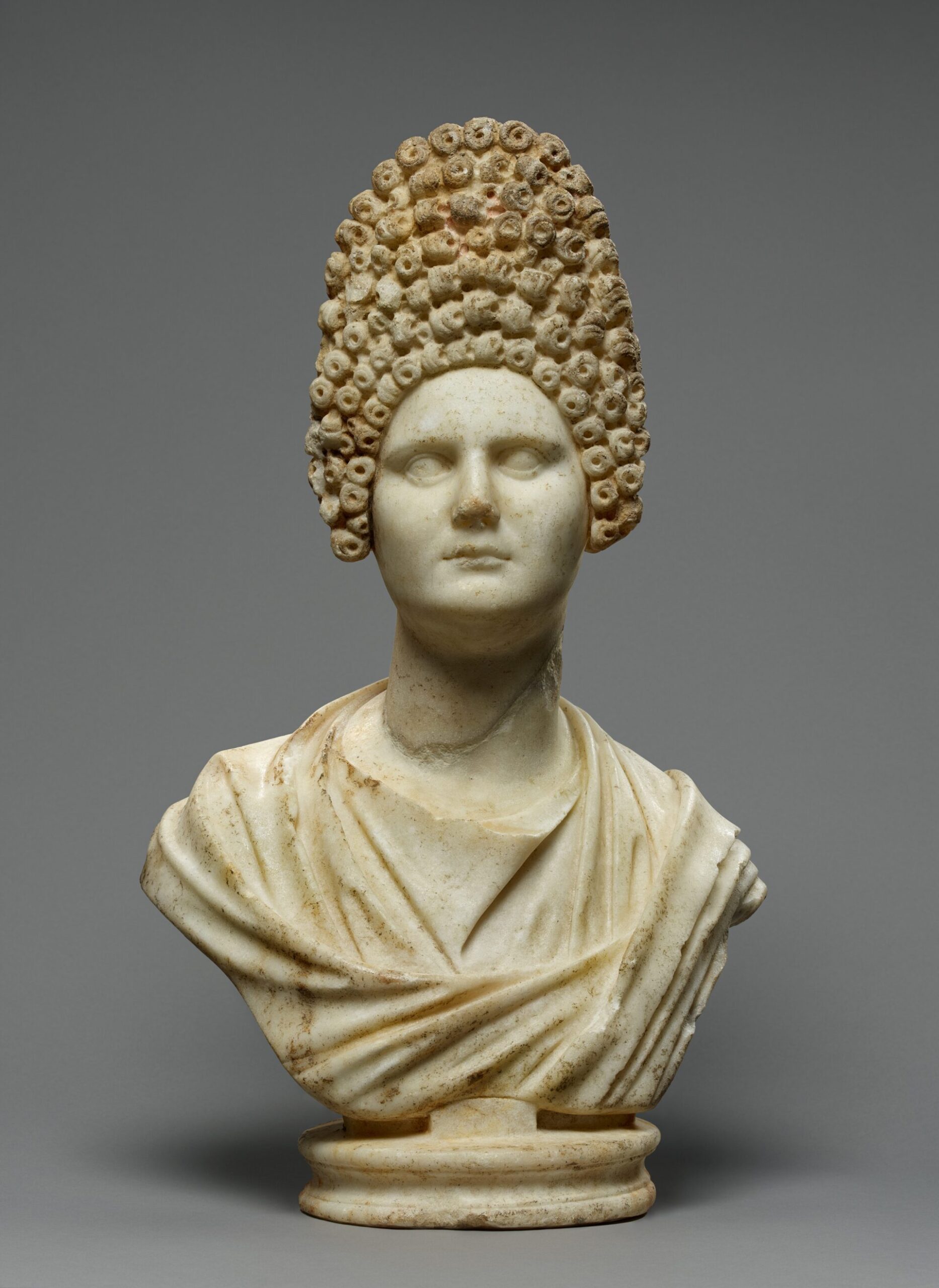 Marble bust of a woman with an elaborate, tall hairstyle of tight curls and draped clothing, set before a plain gray background.
