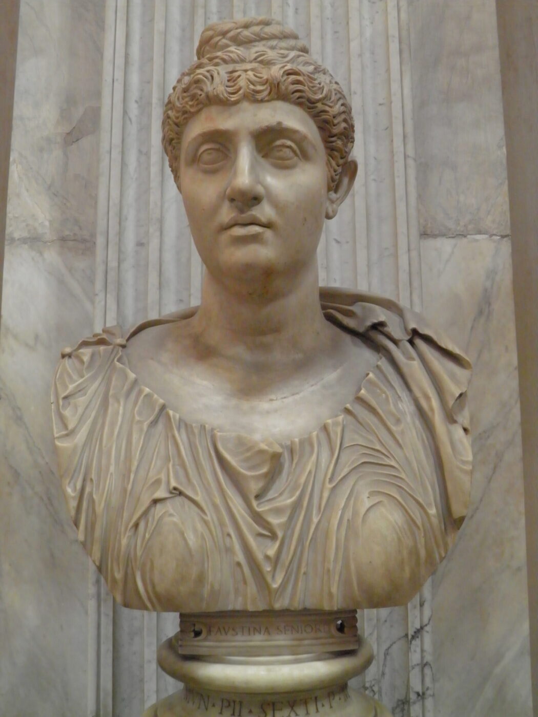 Bust of a woman with wavy hair up, in detailed drapery before marble, classical style, mounted on pedestal.