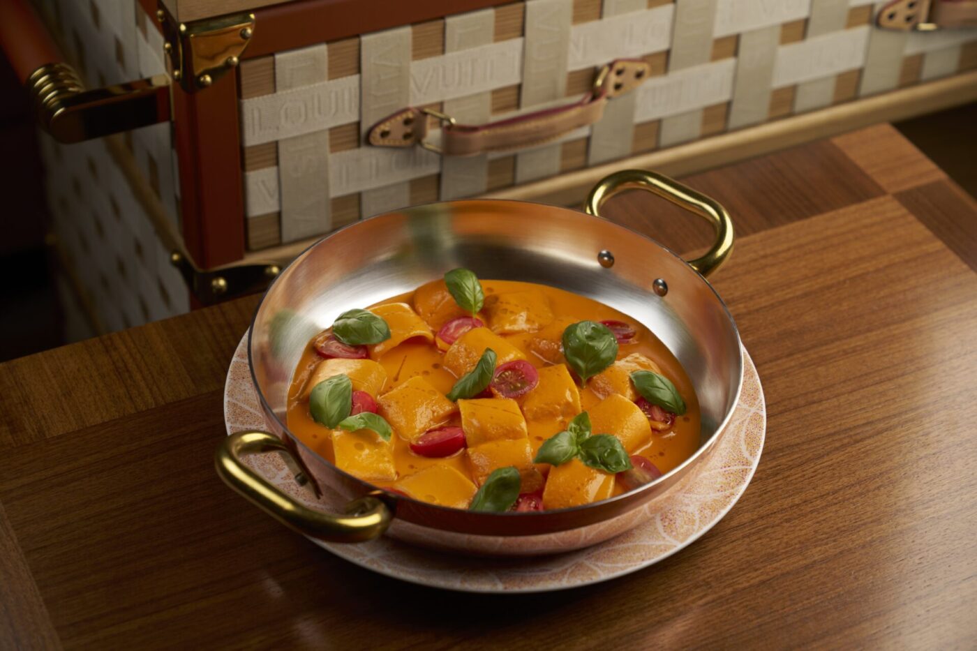 A copper pan with gold handles holds ravioli in orange sauce, topped with cherry tomatoes and basil, on a wooden table by a fancy case.
