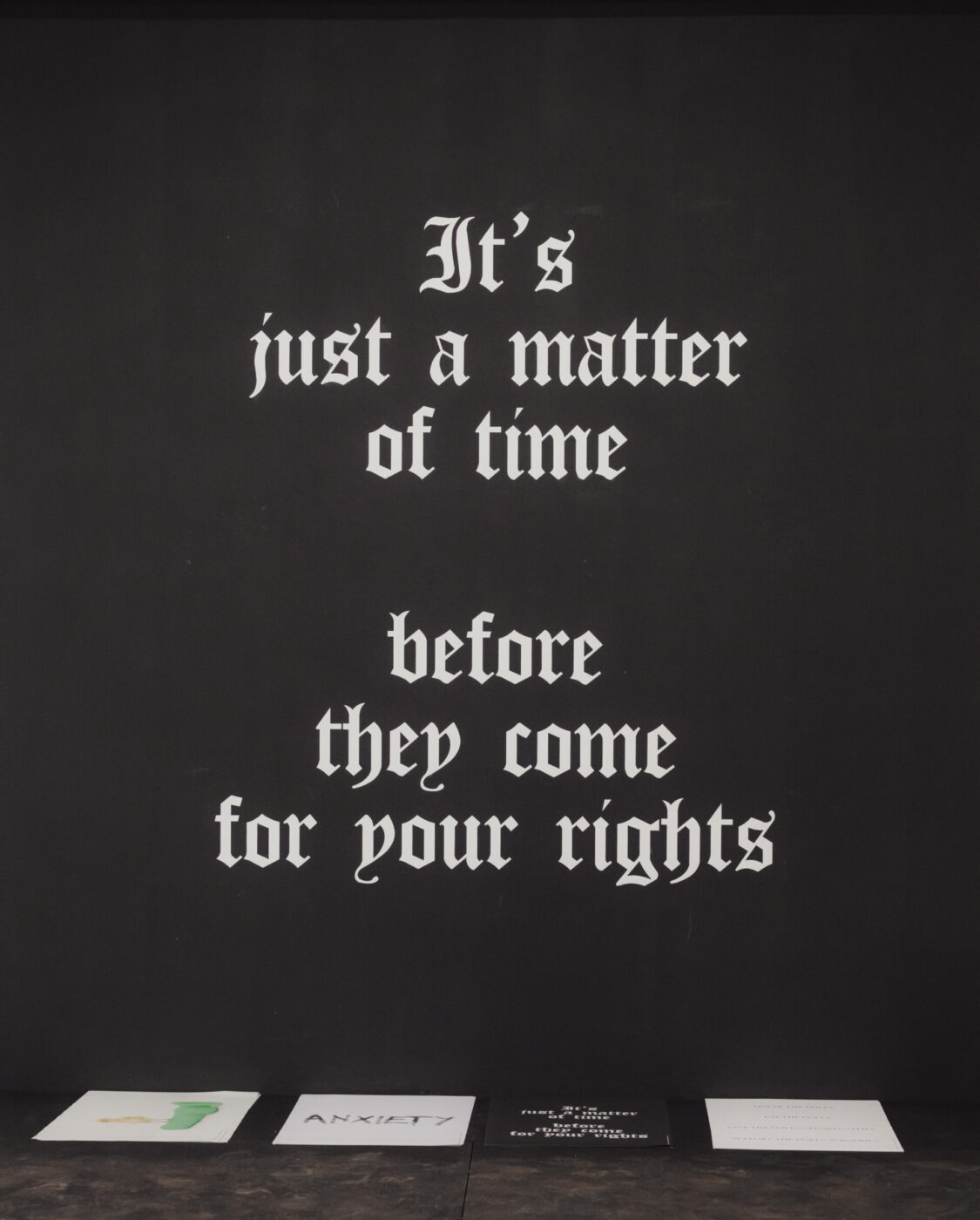 White gothic-style text on a black wall reads, “It’s just a matter of time before they come for your rights,” papers below on dark surface.