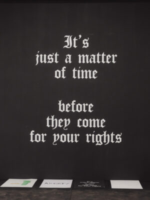 White gothic-style text on a black wall reads, “It’s just a matter of time before they come for your rights,” papers below on dark surface.