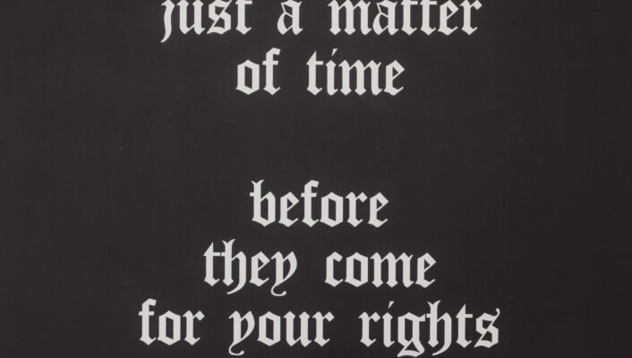 White gothic-style text on a black wall reads, “It’s just a matter of time before they come for your rights,” papers below on dark surface.