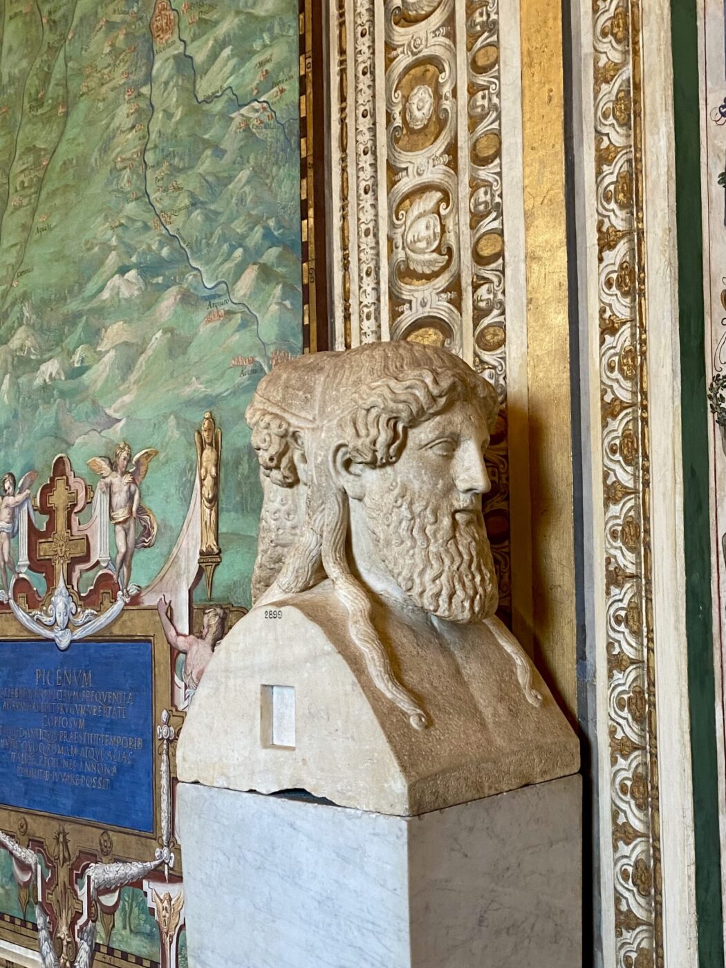 A stone bust of Janus with two faces sits on a marble pedestal in an ornate gallery, surrounded by gilded frames and wall art.