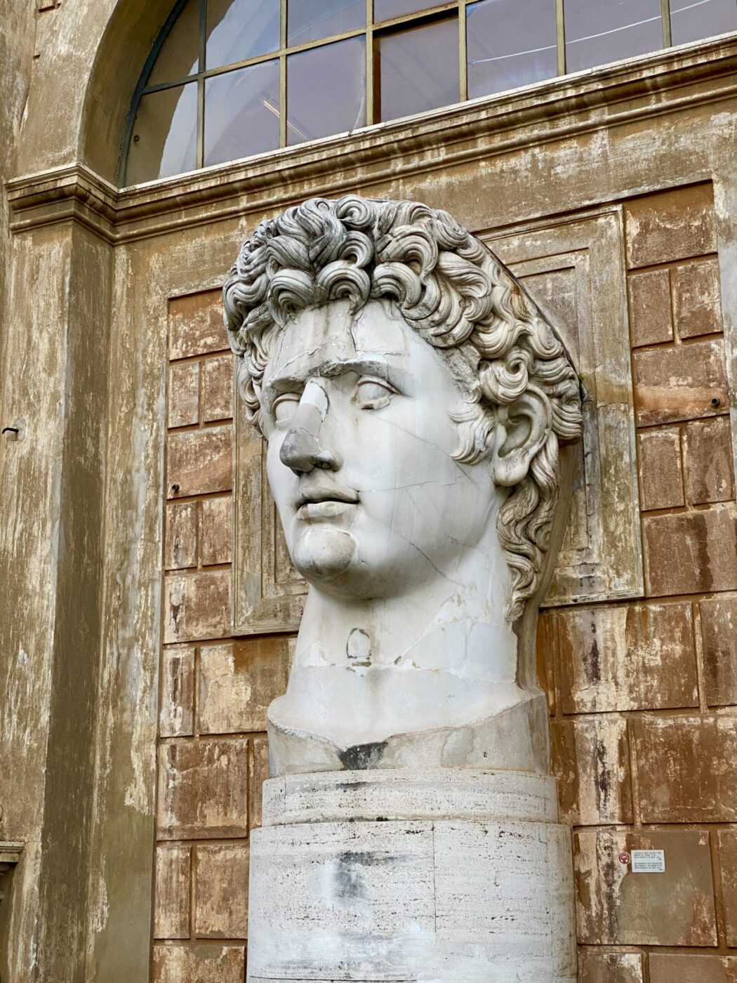 A large ancient marble statue head with a broken nose sits outside a worn stone building featuring arched windows.