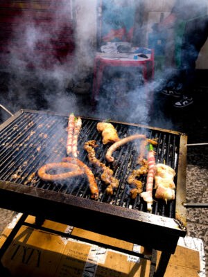 Una griglia fumante cuoce carni assortite, salsicce e spiedini. Vicino, una persona è in piedi accanto a uno sgabello rosso e a un tavolo coperto di utensili all'aperto.
