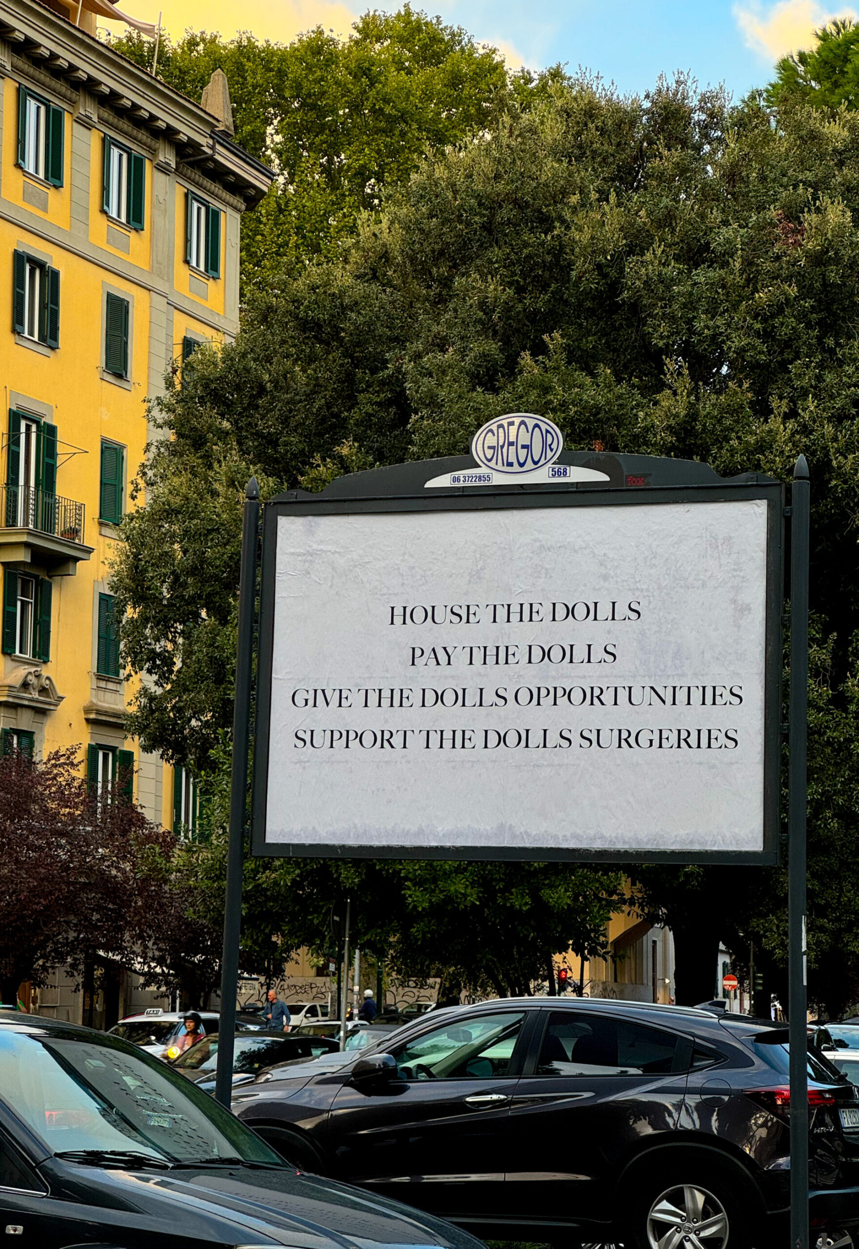 A large street sign says: HOUSE THE DOLLS PAY THE DOLLS GIVE THE DOLLS OPPORTUNITIES SUPPORT THE DOLLS SURGERIES; cars, trees, city.
