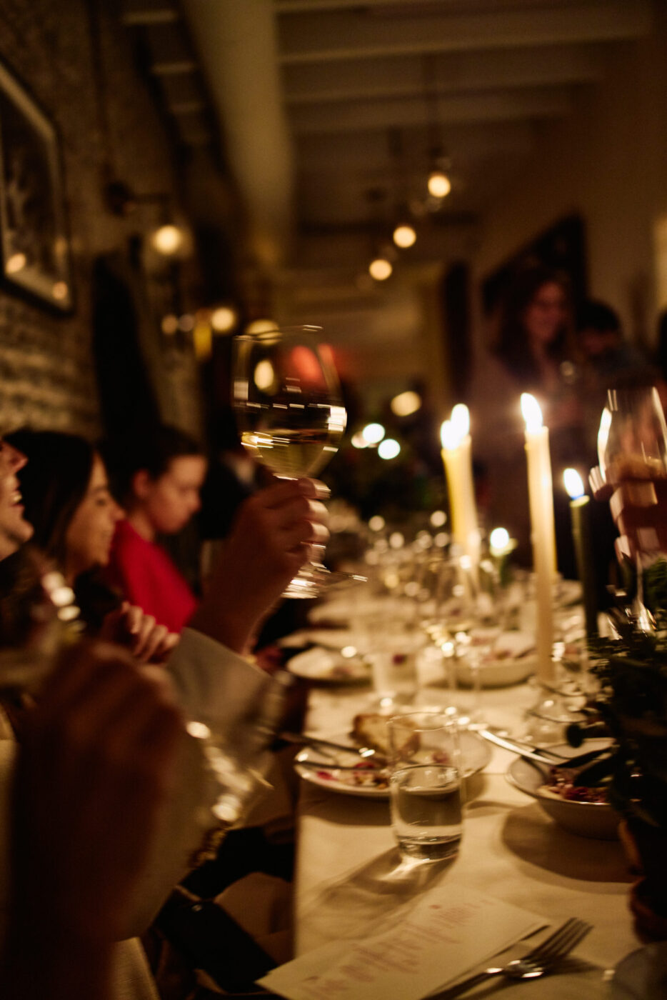 A group of people toast at an elegant, warmly lit dinner table filled with food and candles, creating a festive, intimate mood.