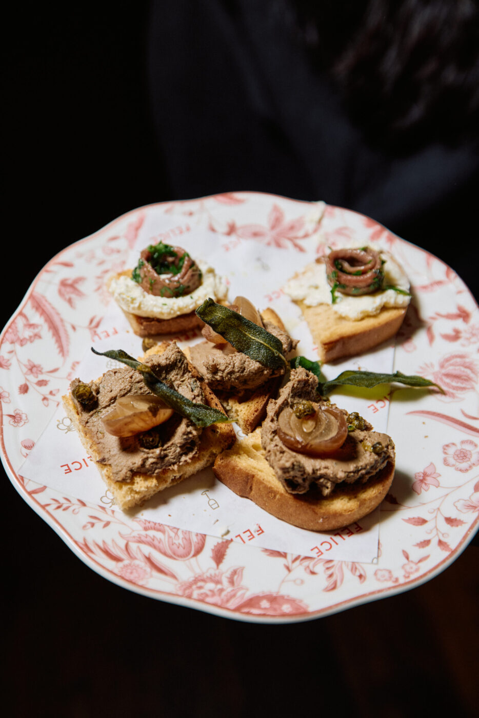 A floral plate with four gourmet toasts, each topped with spreads, herbs, and garnishes against a dark background.