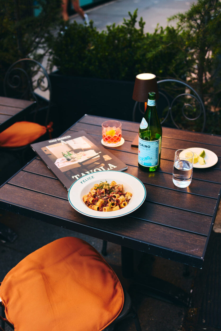 A pasta plate, lemon water, sparkling water, cocktail, and menu on a dark table; orange chairs and green plants in background.