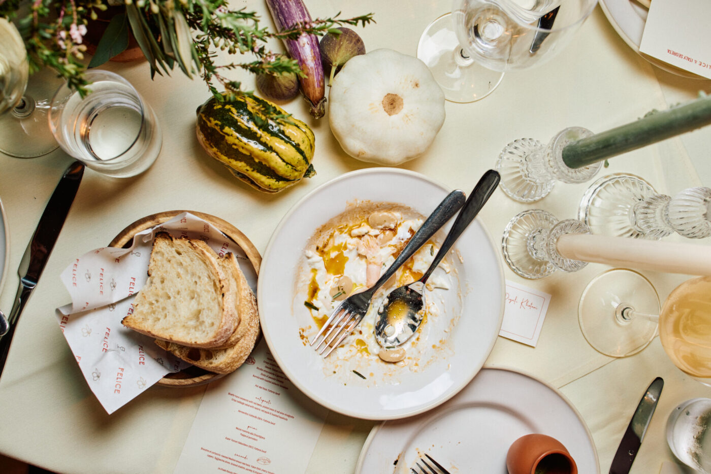 A dinner table with a half-eaten plate, fork, knife, bread bowl, gourds, glassware, candles, and flowers on a cream tablecloth.