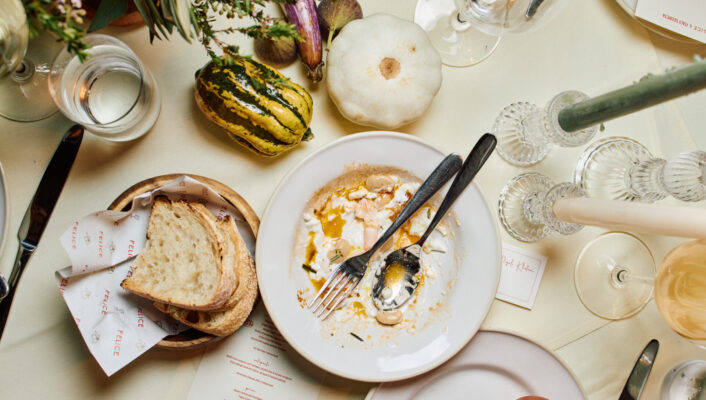 A dinner table with a half-eaten plate, fork, knife, bread bowl, gourds, glassware, candles, and flowers on a cream tablecloth.