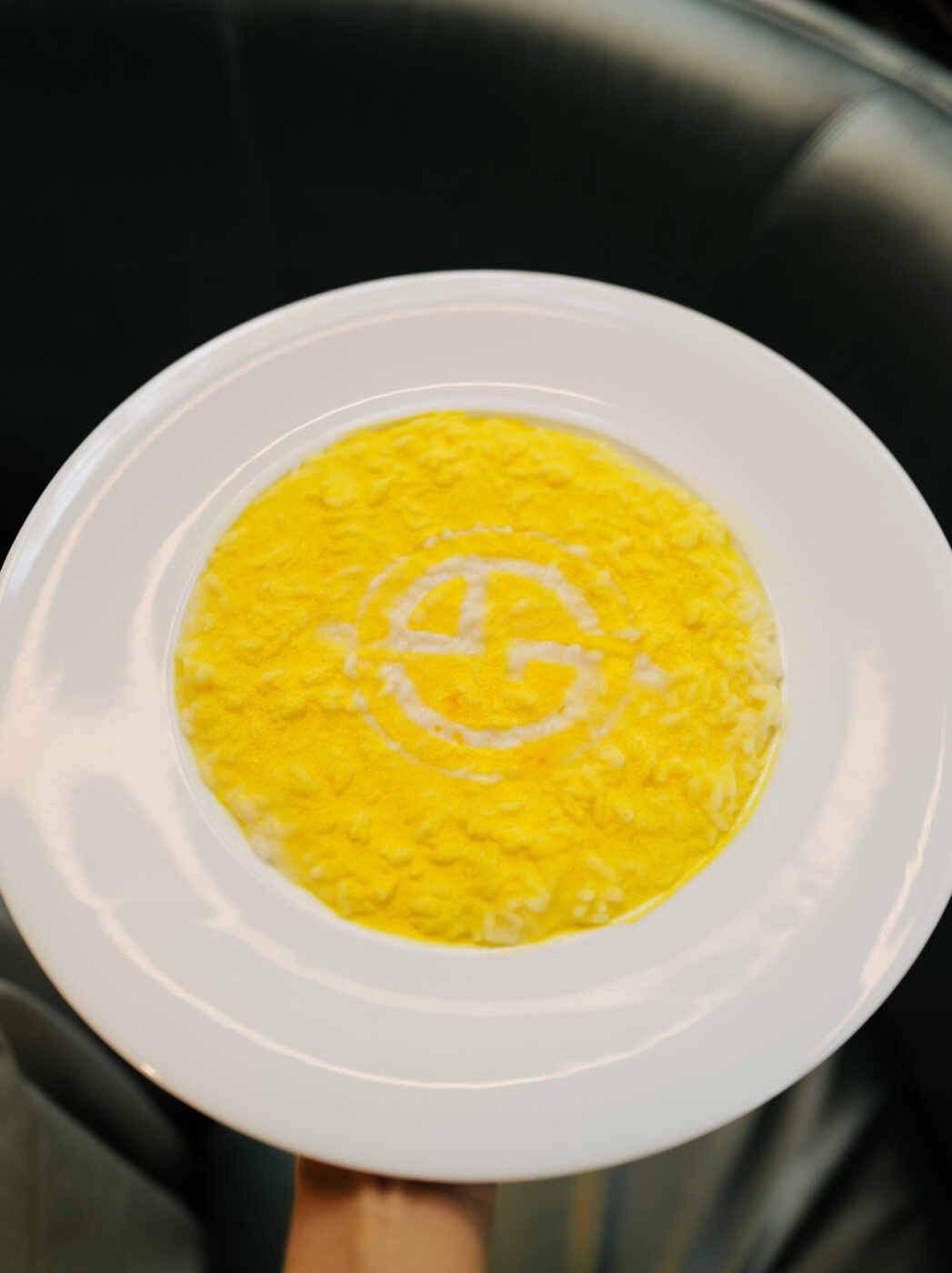 A white plate with creamy yellow risotto, topped with a stylized white “G” design, seen from above on a dark background.