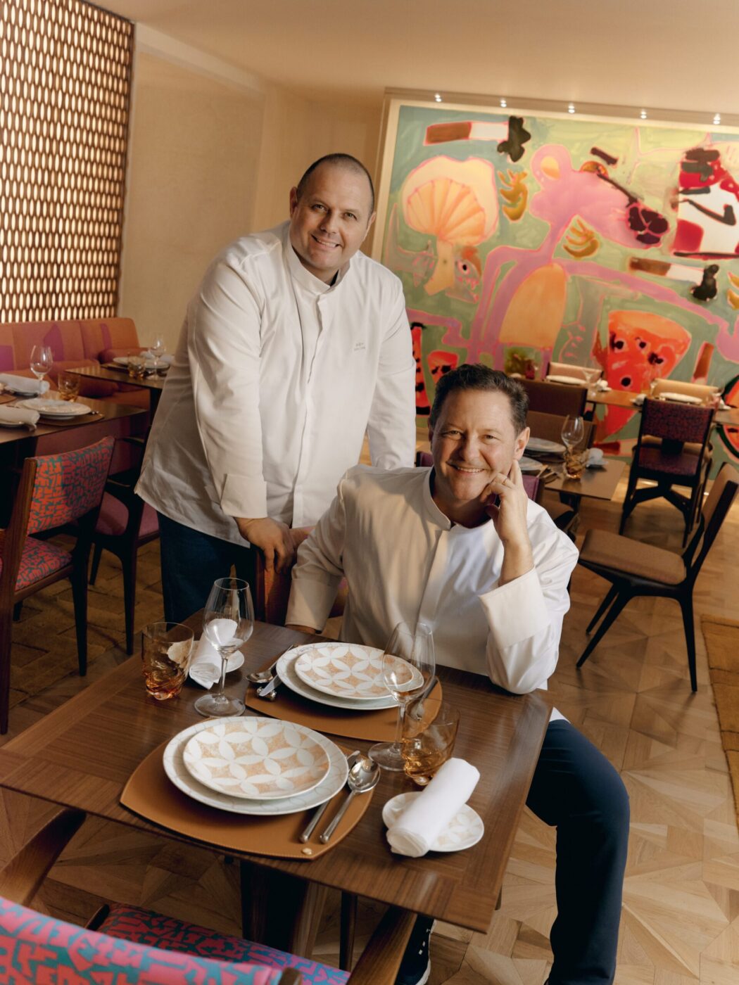 Two chefs in white jackets smile in a chic restaurant with colorful art, set tables, and modern decor; one sits as the other stands.