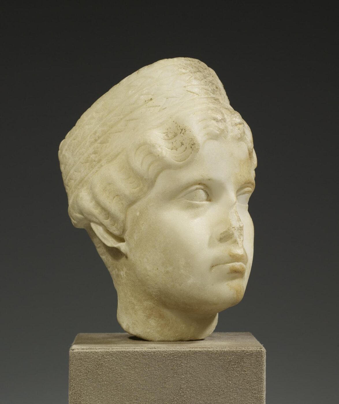 A marble sculpture of a young head with wavy hair and a cap, set on a stone pedestal against a dark plain background.
