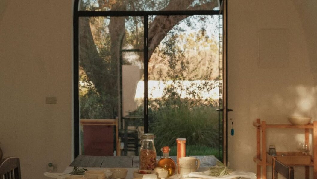 A sunlit breakfast table with pastries, bread, fruit, and cheese in a cozy room; arched window shows trees and greenery.