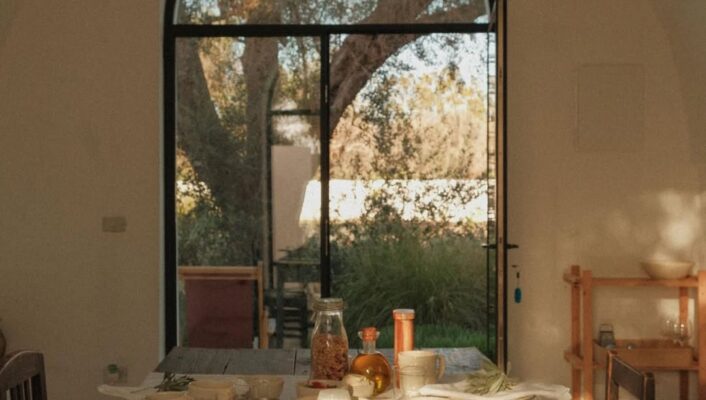 A sunlit breakfast table with pastries, bread, fruit, and cheese in a cozy room; arched window shows trees and greenery.