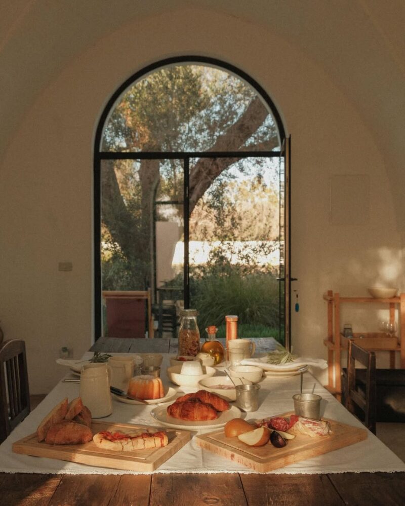 Una sala da pranzo illuminata dal sole con un tavolo per la colazione con dolci, frutta, formaggio e caffè. La finestra ad arco mostra un giardino verdeggiante.