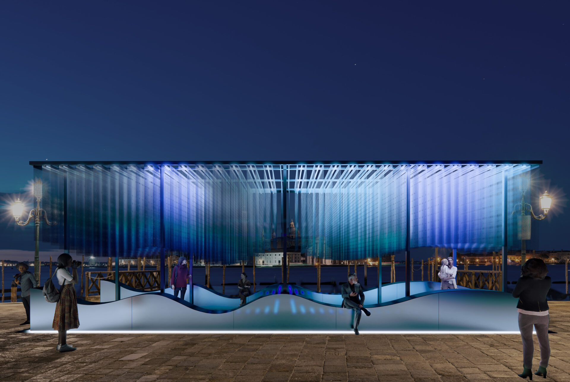 A nighttime art installation with wavy blue and white hangings; people stroll nearby, with city lights and water in the background.