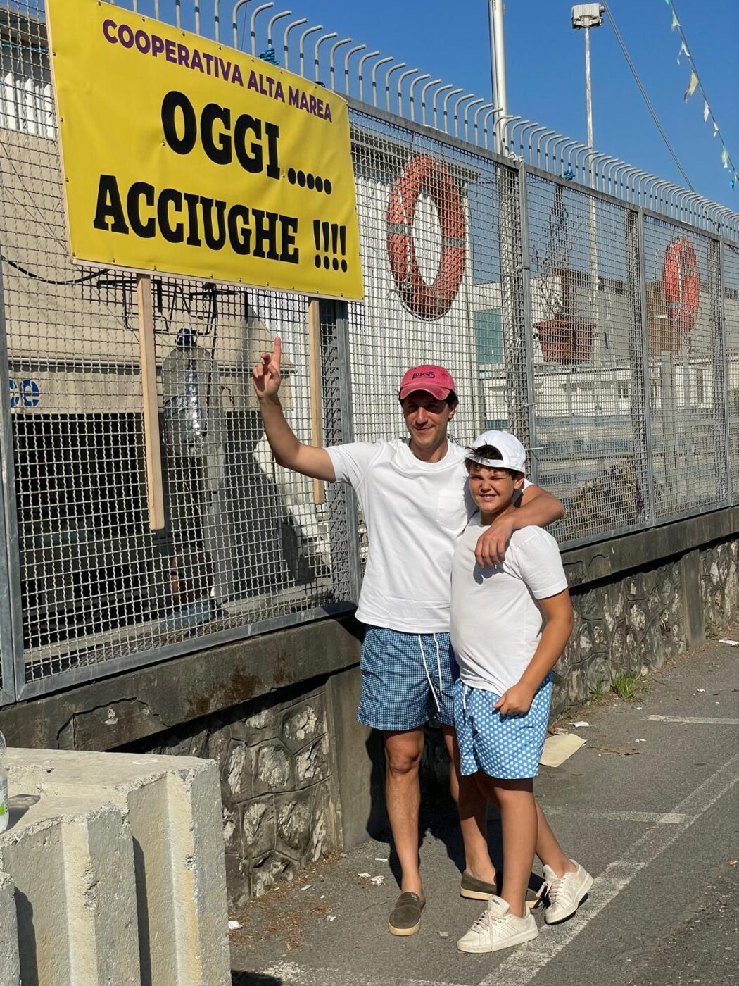 [OGGI… ACCIUGHE !!!] - Two people in summer outfits and hats smile under a yellow "OGGI… ACCIUGHE !!!" sign by a fenced area.