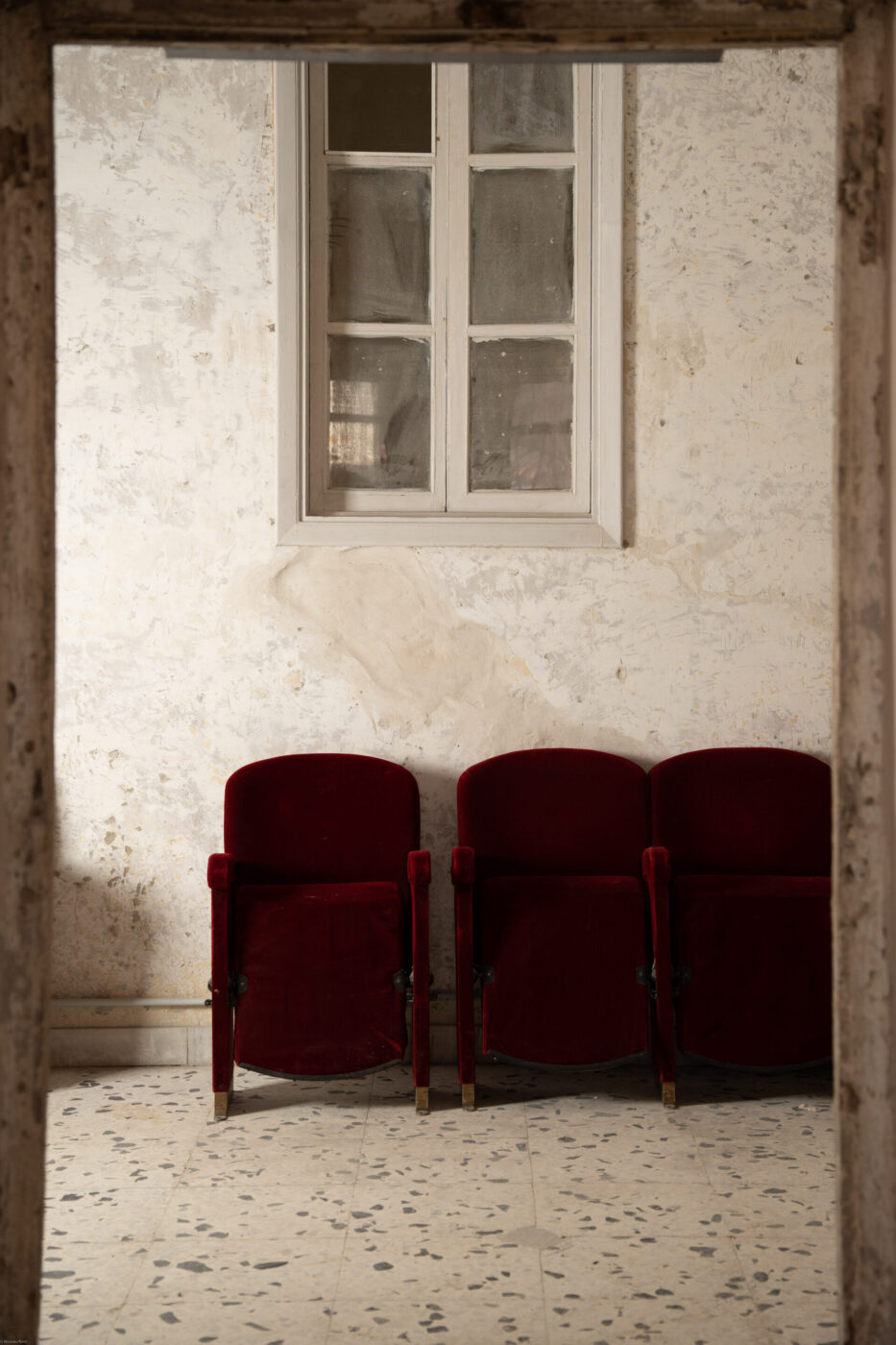Three red velvet theater seats in a rustic room face a worn wall and closed window, with a speckled tile floor beneath them.