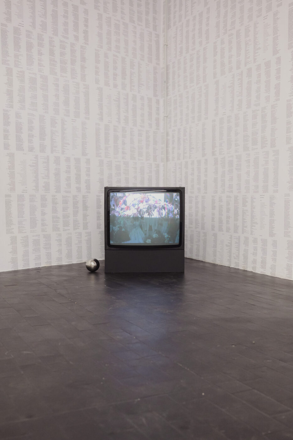A vintage TV showing a crowded scene sits on a dark tile floor; microphone nearby, walls lined with dense lists or names.