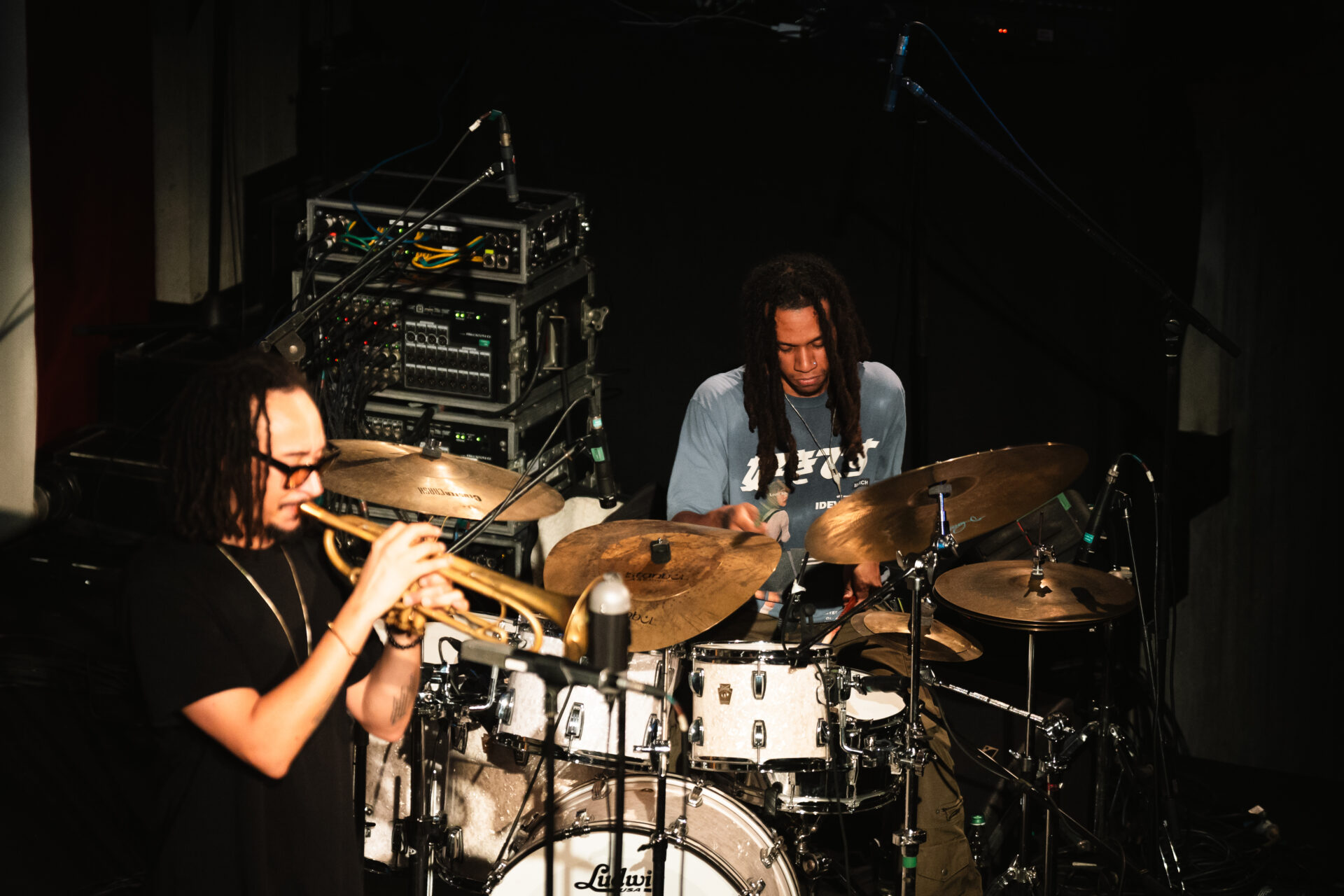 A trumpet player and a drummer with dreadlocks perform on stage; the trumpet is in front, the drummer at a white drum kit behind.