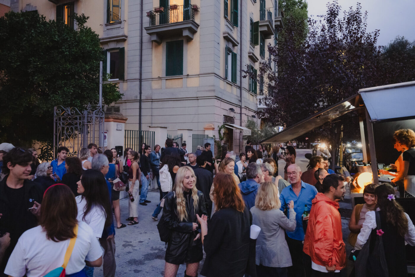 A crowd gathers by a food stall on a city street at dusk, mingling near an apartment as streetlights and trees line the background.