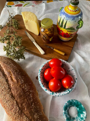 A rustic spread: sesame bread, red tomatoes, cheese, figs, herbs, olive oil bottle, and knife on a board over a white cloth.