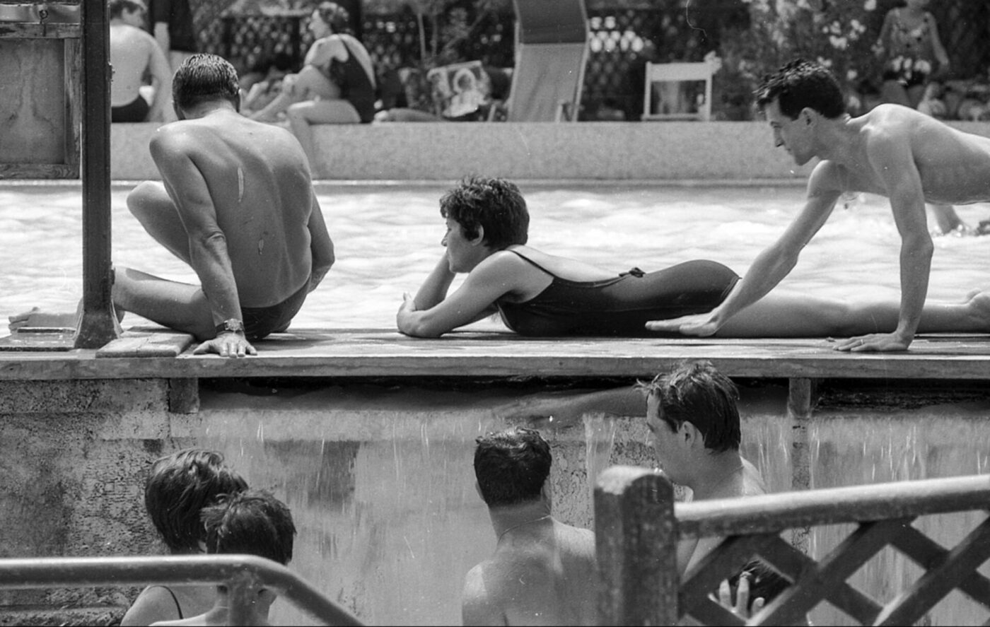 Black and white photo of poolside relaxation; two men and a woman lounge at the edge, others stand in water, more in background.