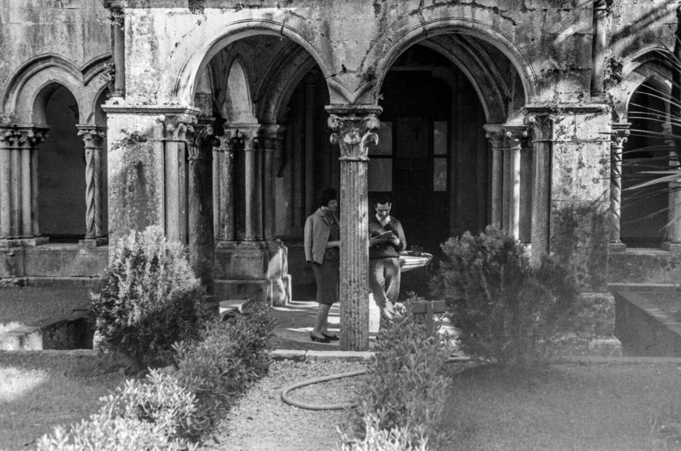 Two people talk by a stone pillar in a historic cloister garden, arched walkways and greenery, sunlight casting shadows on old walls.
