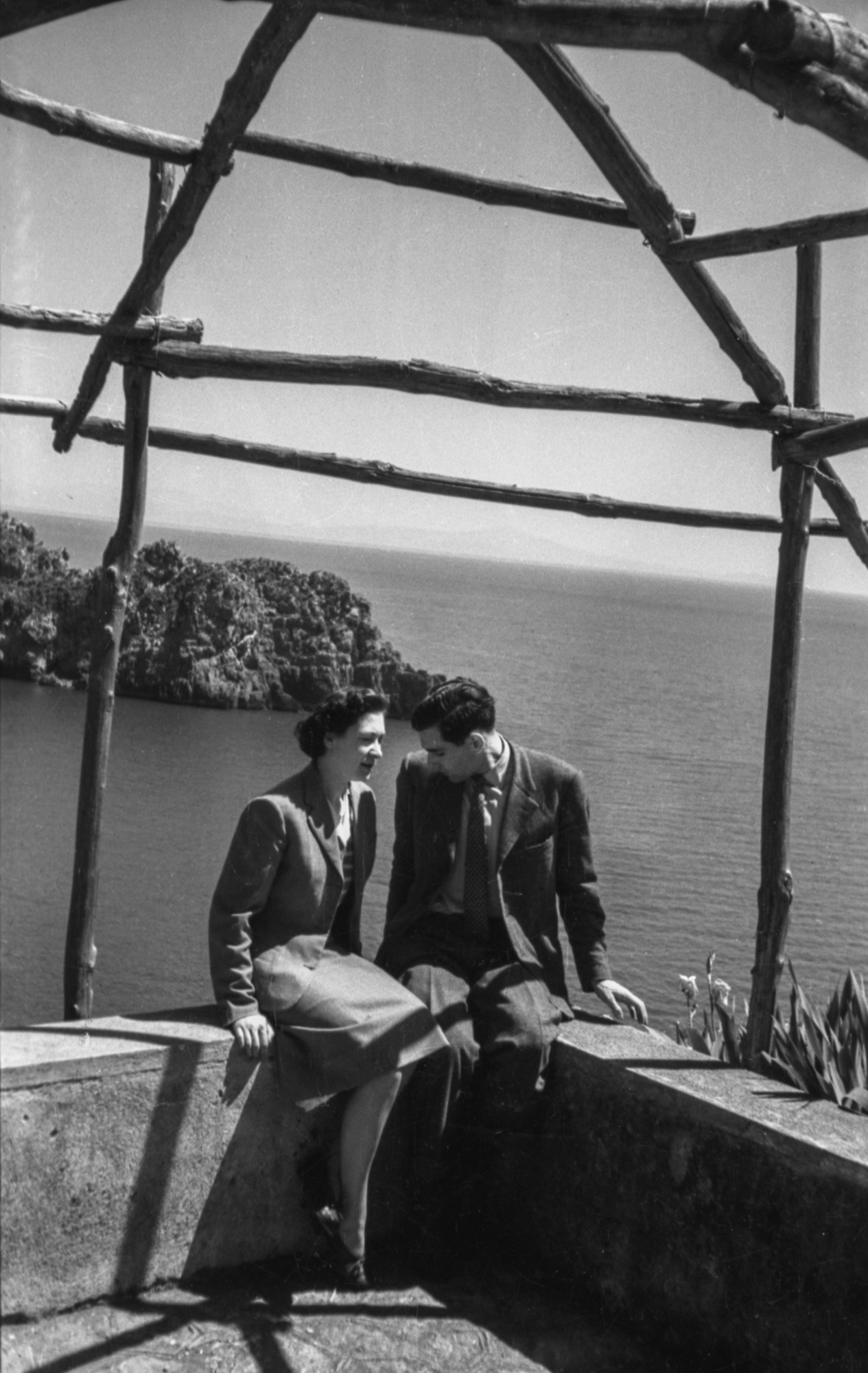 A woman and man in vintage clothes sit on a stone ledge under a wooden frame, cliffs and sea behind them on a sunny day.