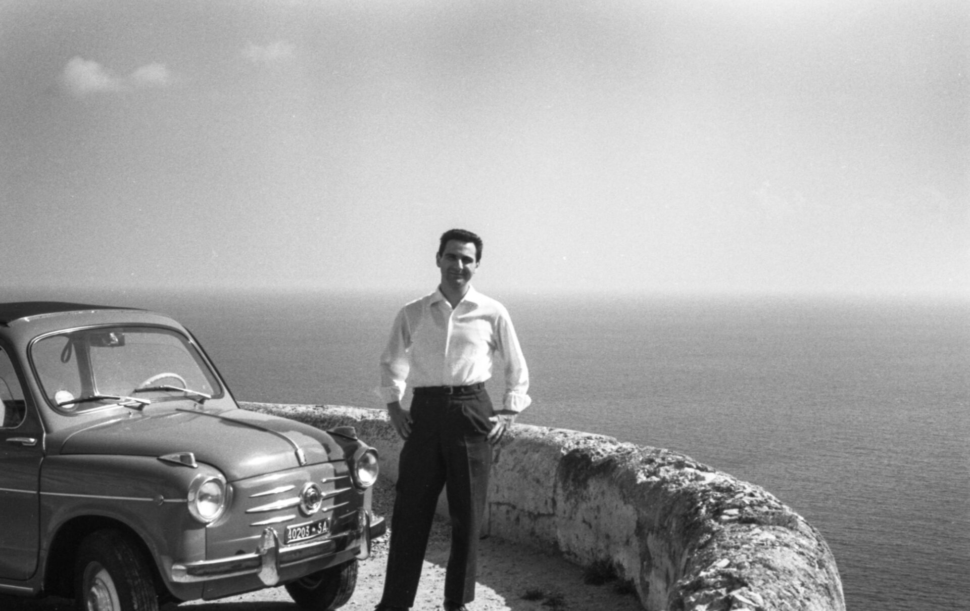 A man in a white shirt and dark pants stands by a vintage car on a stone overlook, sea and horizon behind. Black and white photo.