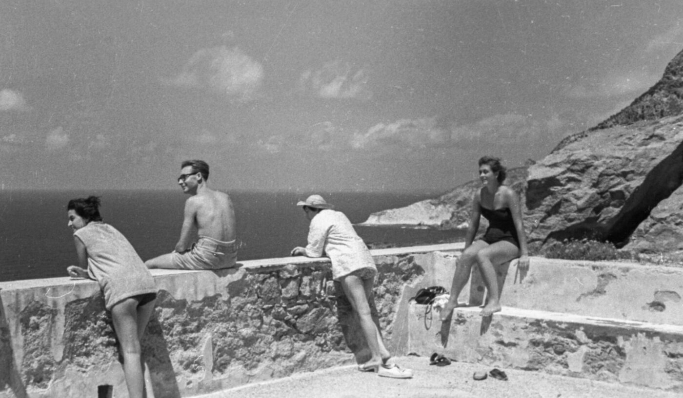 Four people in swimwear sit and lean on a stone wall by the sea, facing rocky cliffs; two women left, man and woman right.