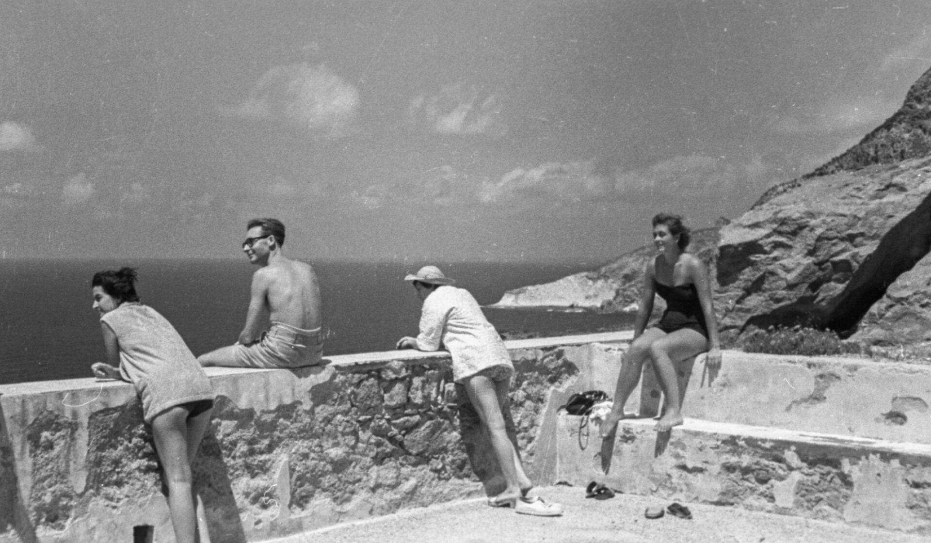 Four people in swimwear sit and lean on a stone wall by the sea, facing rocky cliffs; two women left, man and woman right.