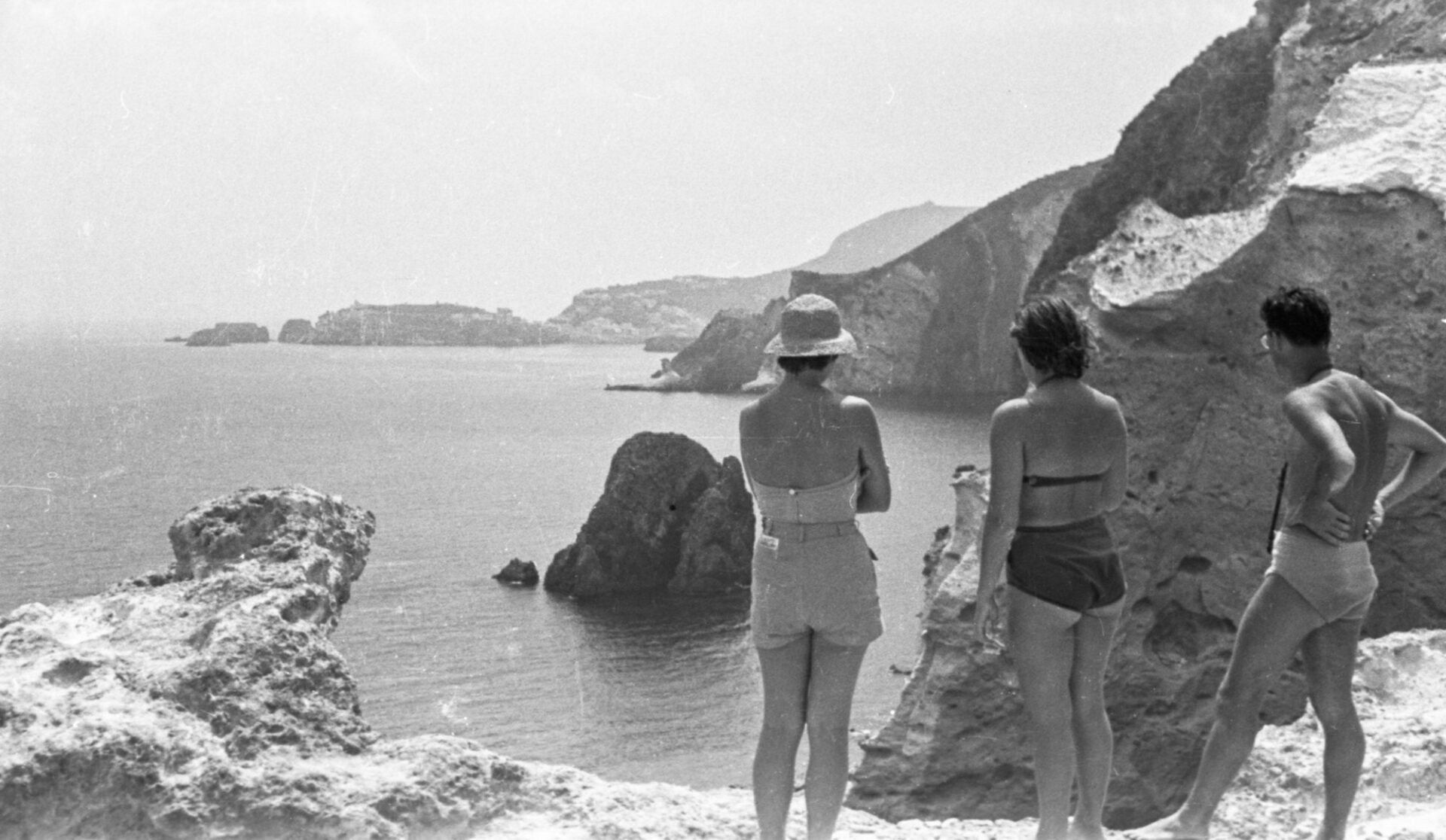 Three people in swimwear stand on rocky cliffs above a calm sea with distant hills and rock formations under a hazy sky.