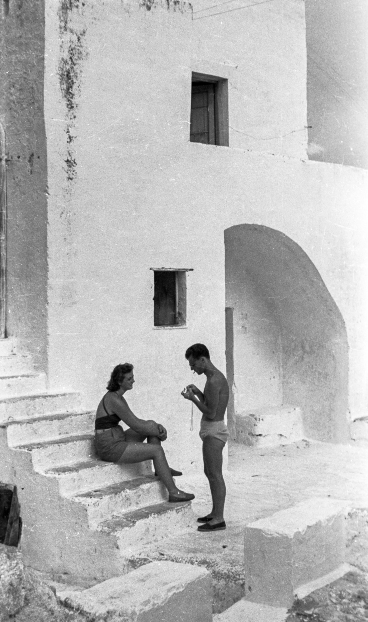 A woman sits on white stone steps as a man stands nearby in swimwear outside a sunlit, whitewashed building with arches.