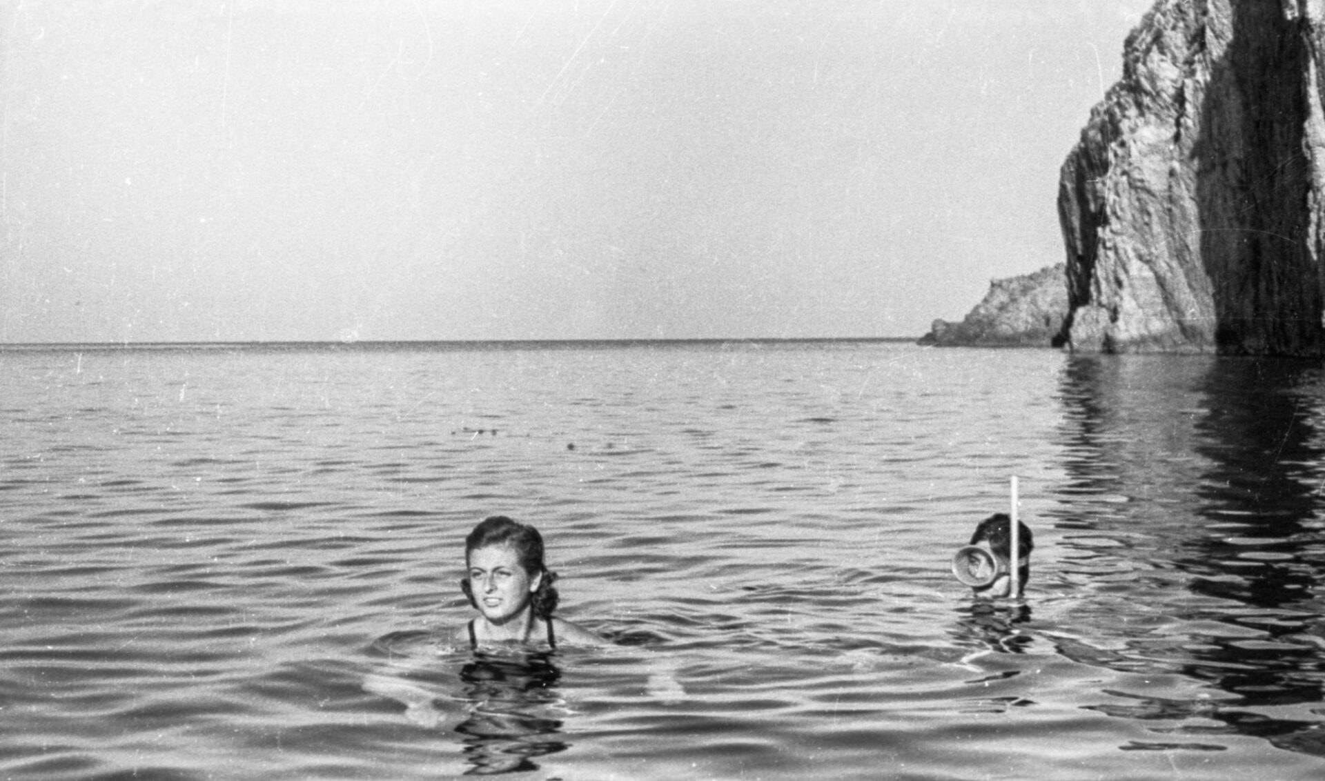 Two people swim in calm ocean water by a rocky cliff; one faces forward, the other wears a snorkel. Black and white, open sky.