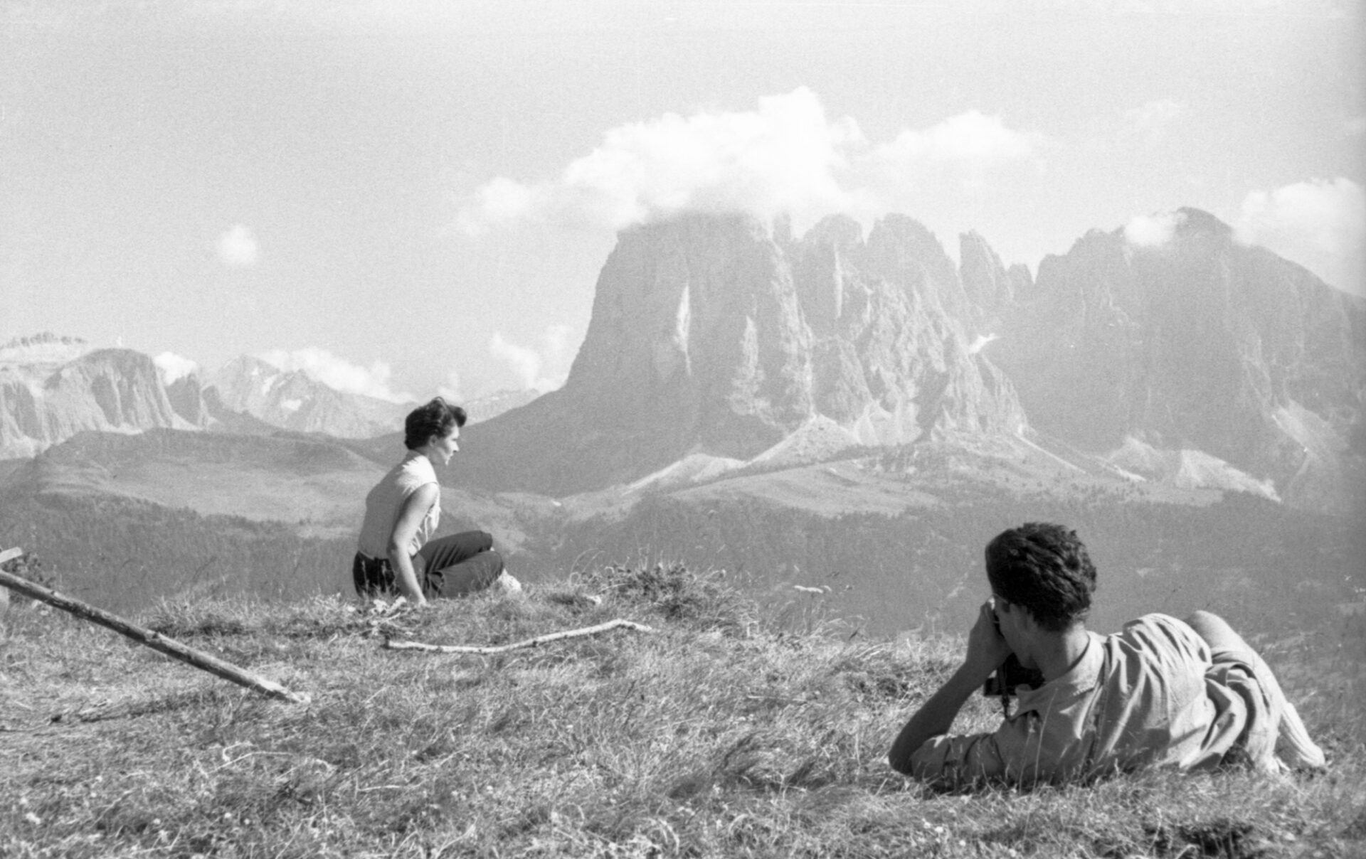 Two people relax on a grassy hillside, one sitting and one lying down, gazing at rugged mountains under a tranquil partly cloudy sky.