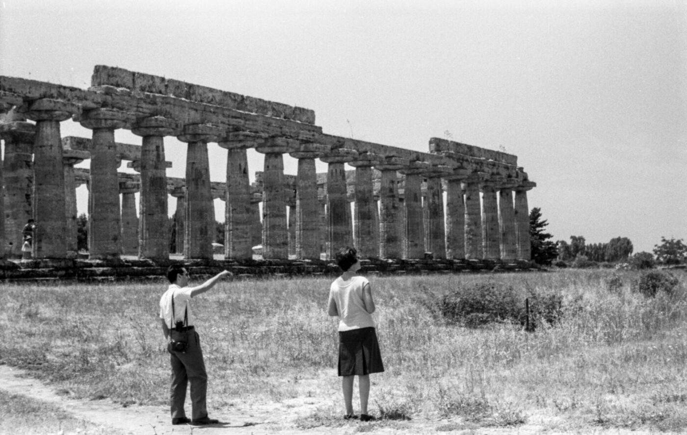 Two people in a grassy field by ancient stone columns of a temple, one pointing at the ruins under a clear sky.