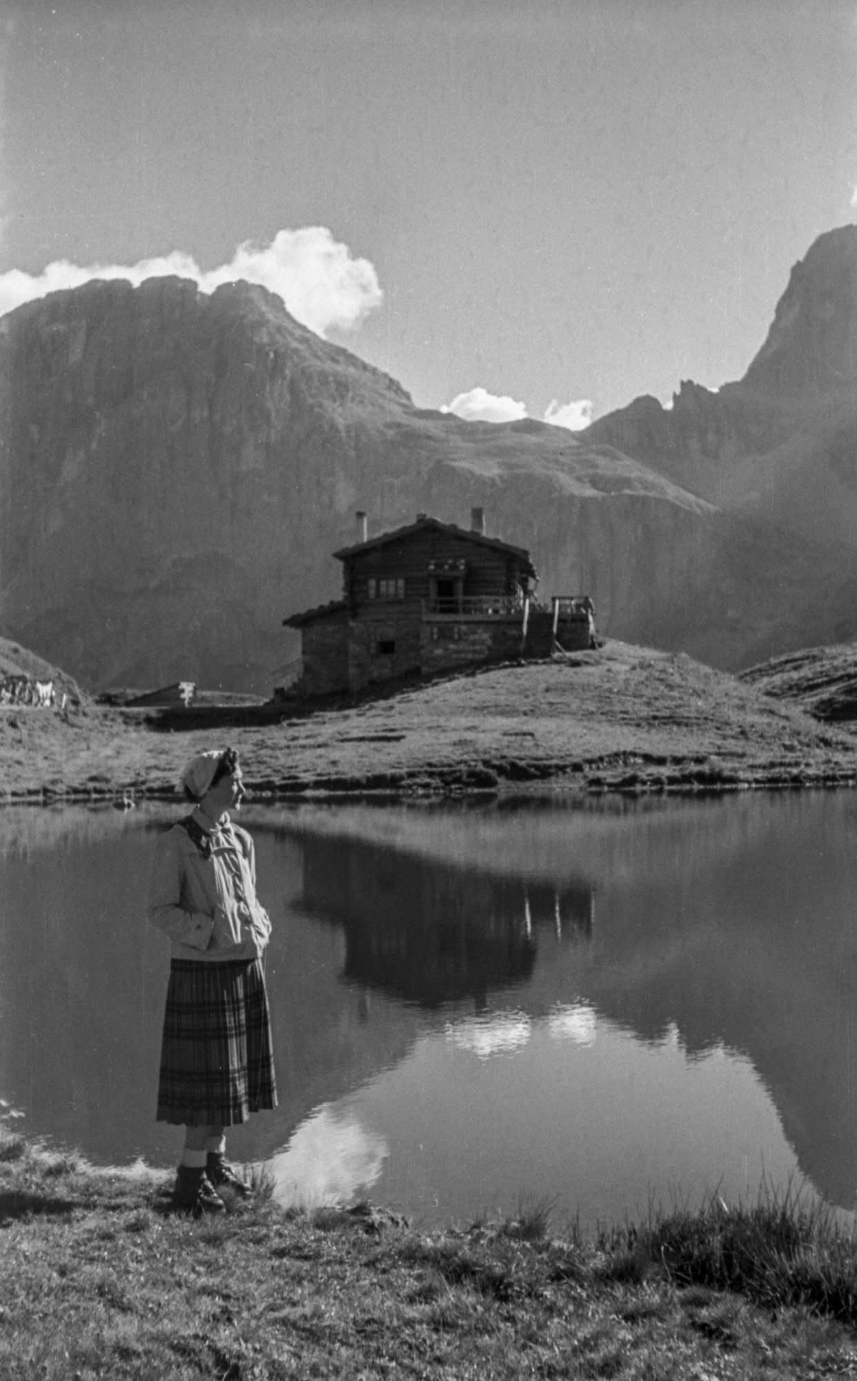 A person in a skirt, jacket, and headscarf stands by a lake, gazing at a cabin. Mountains and clouds are mirrored in the calm water.