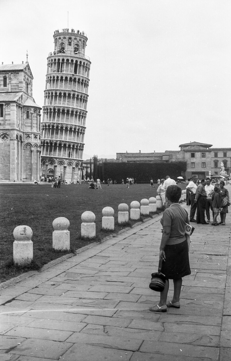 A black-and-white photo shows a woman on a pathway gazing at the Leaning Tower of Pisa, with tourists and old buildings behind her.