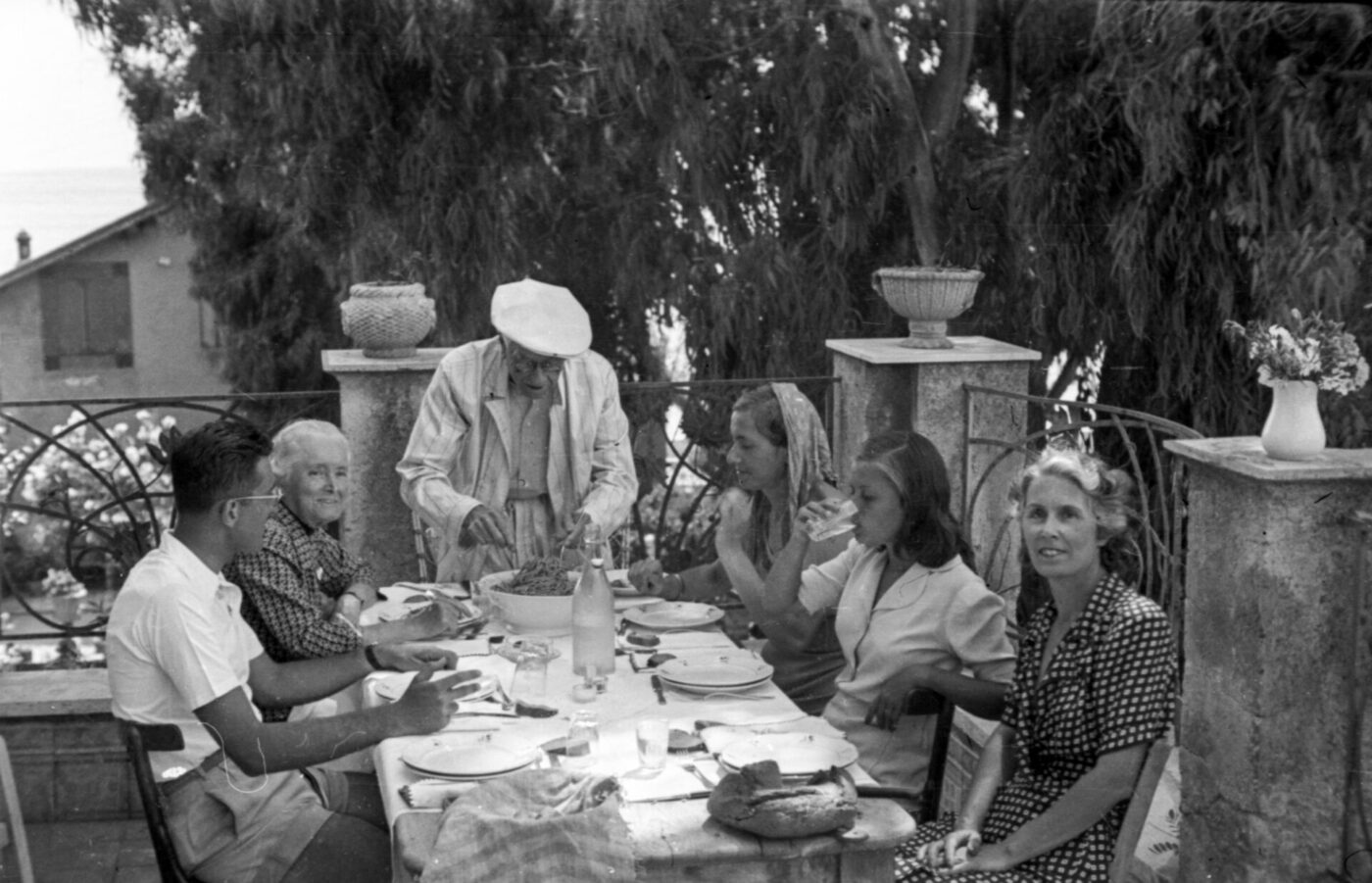 Six people gather at a shaded outdoor table, eating and talking, with stone railings and potted plants around them.