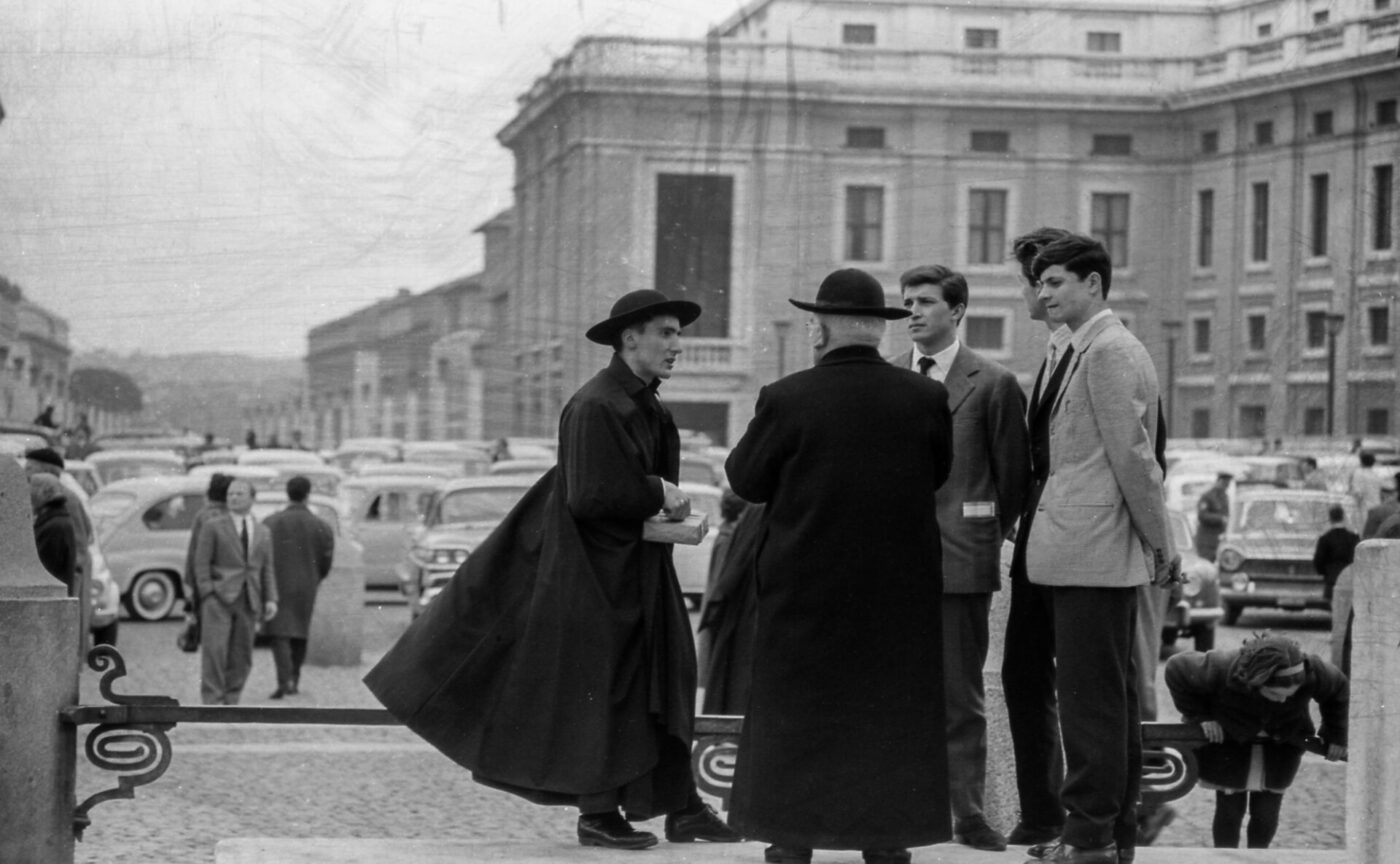 Two priests talk with two young men on a busy mid-century street; cars, historic buildings, pedestrians, and a child tying shoes.