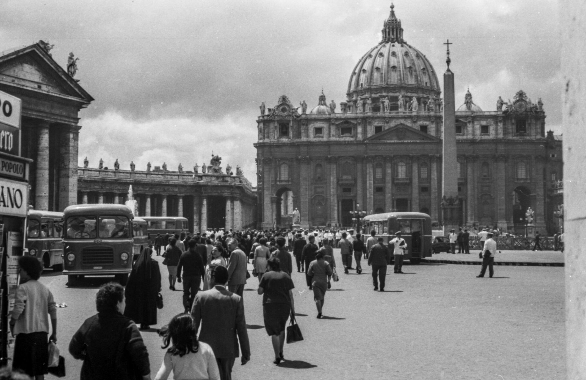: Black-and-white photo of a crowd heading to St. Peter’s Basilica, buses parked nearby, large dome and obelisk in the background.