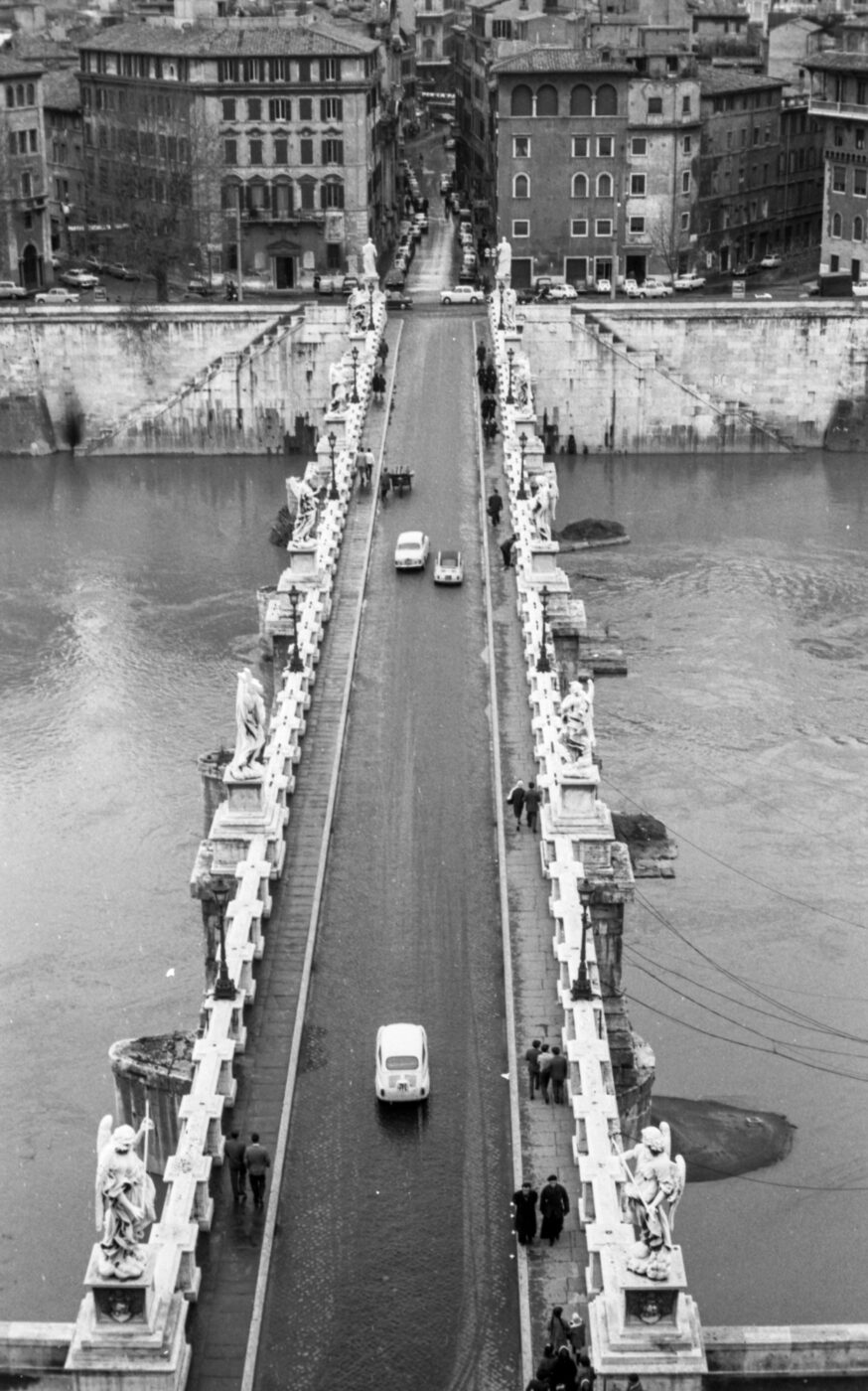 A black-and-white aerial view shows a stone bridge with statues spanning a river in an old city, with cars and people crossing.